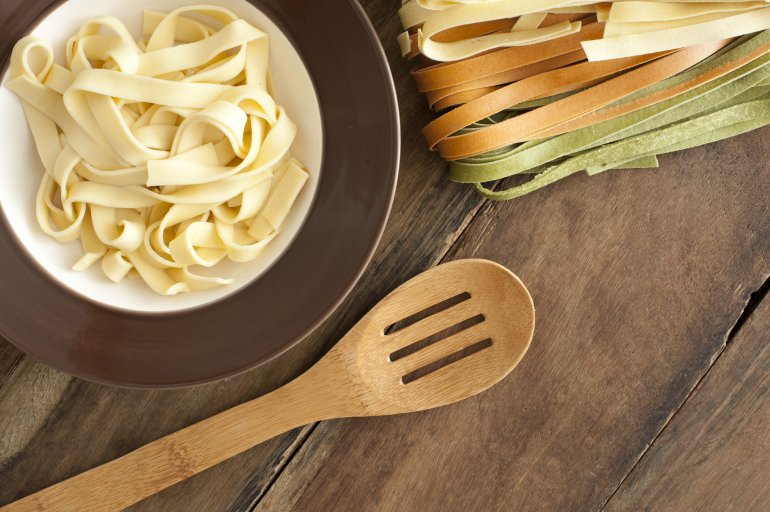 Serving tagliatelle noodles in a small bowl with a wooden straining spoon and assorted varieties of dried tagliatelle pasta alongside, overhead view on a rustic table