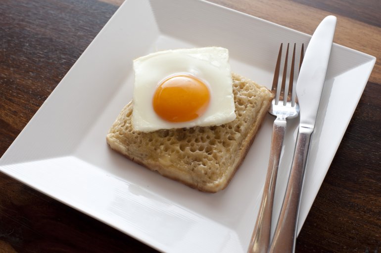 Sunny side up egg cut as square on top of matching shaped fried crumpet patty and plate with fork and knife on top