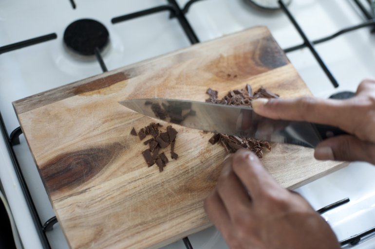 Close up view of the hands of a male chef or cook chopping chocolate for baking on a wooden cutting board on a gas hob