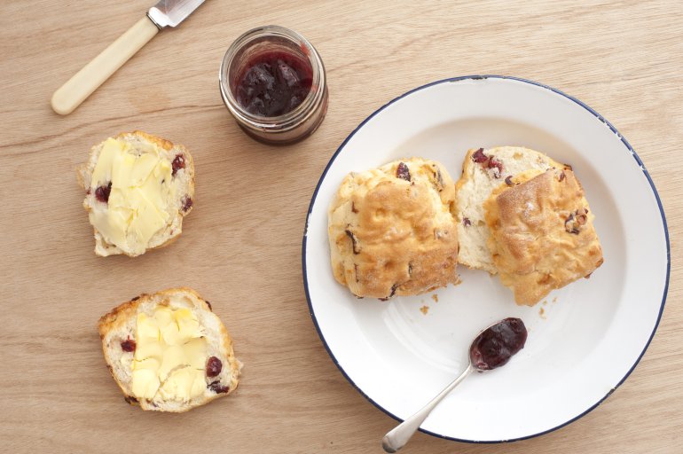 Freshly baked buttered scone or rock cake with raisins and fruit pieces with a jar of strawberry jelly and further cookies on a white plate alongside, overhead view