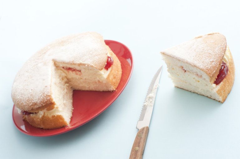 Freshly baked jam sponge cream cake with a generous slice on the side alongside a knife, served on a red plate
