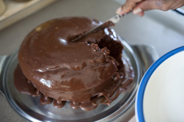 Person cutting a freshly baked mud cake or chocolate sponge dripping with chocolate icing serving a slice onto a side plate