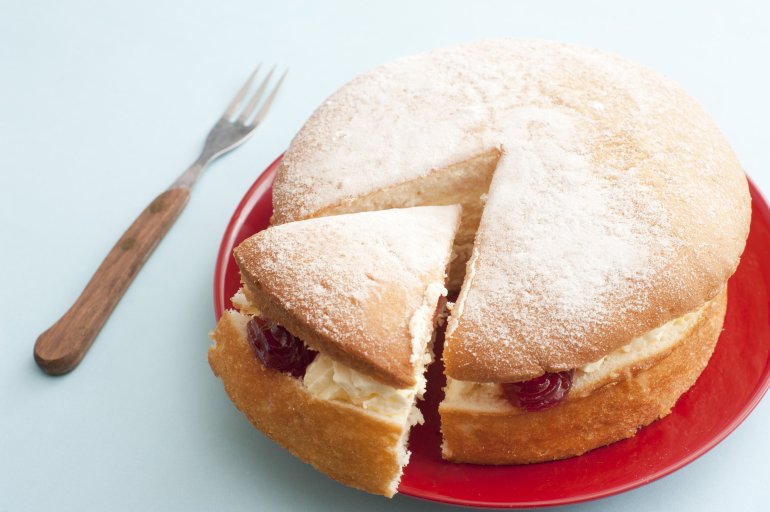 Freshly baked plain vanilla sponge cake filled with cream and strawberry jam with a single slice cut and moved to the side of the red plate , close up high angle view