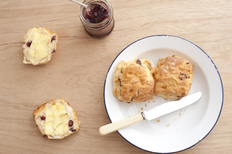 Buttered freshly baked homemade fruit scones served on a plate and wooden table with a jar of berry jam , overhead view