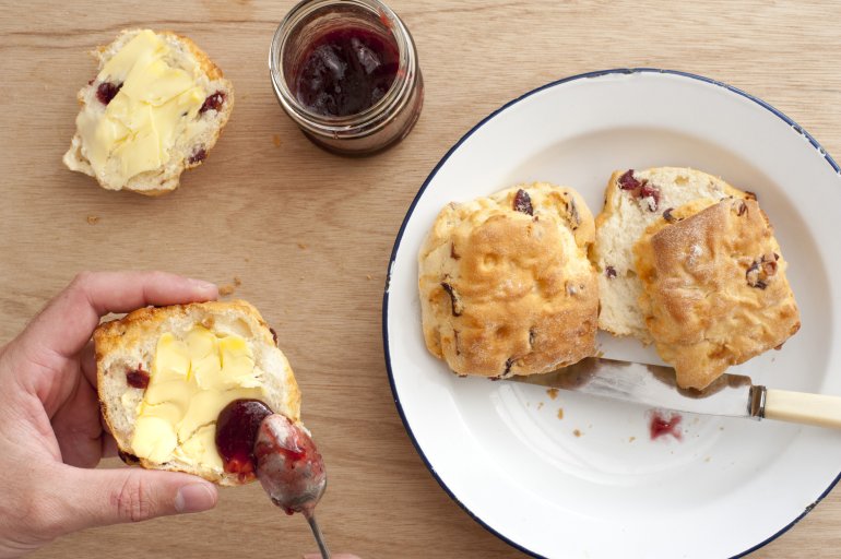 First person perspective of hands spreading strawberry jam on buttered crispy scone biscuits with spoon over plate on light brown wooden table