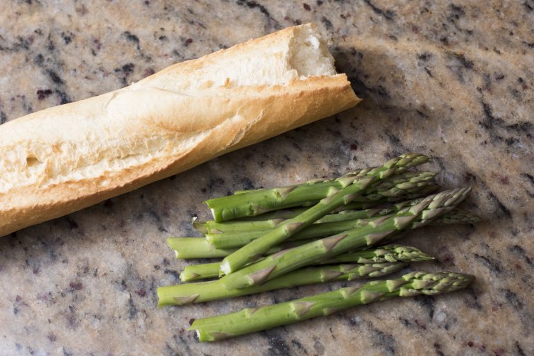 Fresh green asparagus tips and a crusty white baguette on a marble counter top in the kitchen