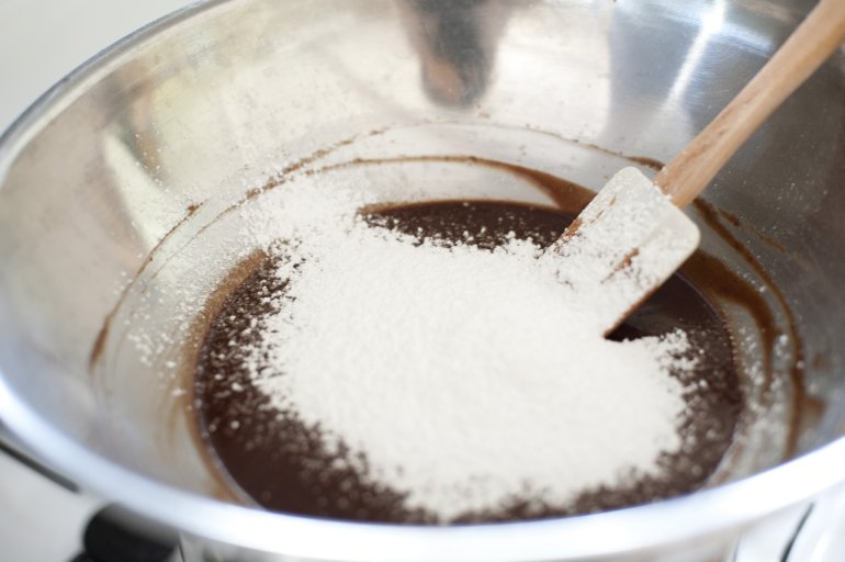 Cook making chocolate icing adding icing sugar to the melted chocolate in a mixing bowl, close up view