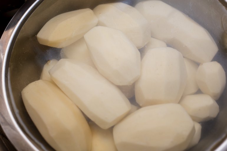 Overhead view of a pile of peeled white potatoes cooking in a deep pot with a clear cover