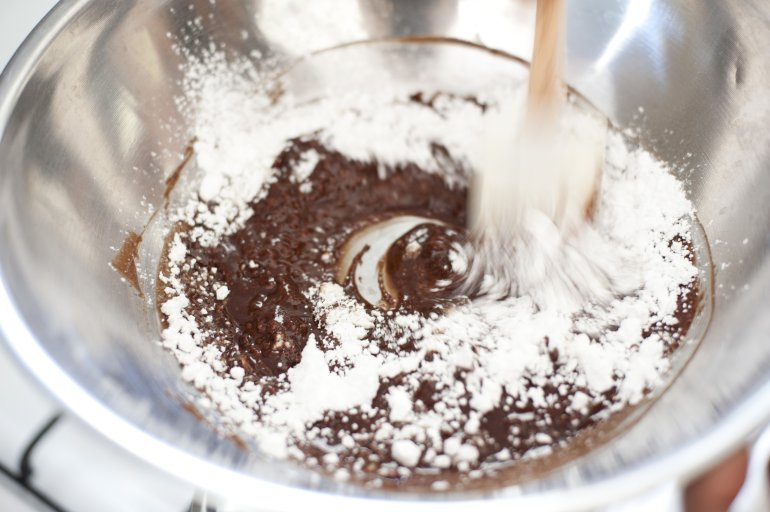 Overhead closeup view of powdered sugar mixing with melted chocolate in a steel bowl