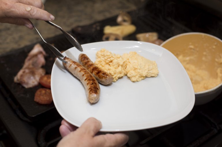 Man preparing hid breakfast serving up to cooked sausages from a grill onto a modern white plate, close up of his hands