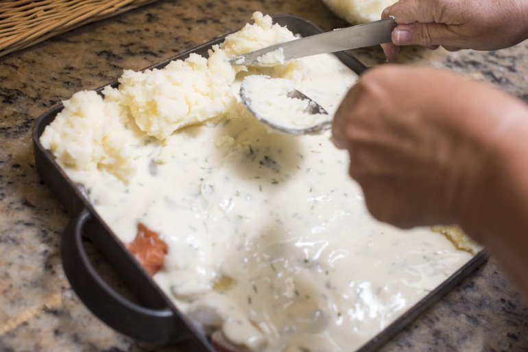 Chef topping a fish pie with mashed potato spooning the mixture onto the top over a creamy sauce, close up view