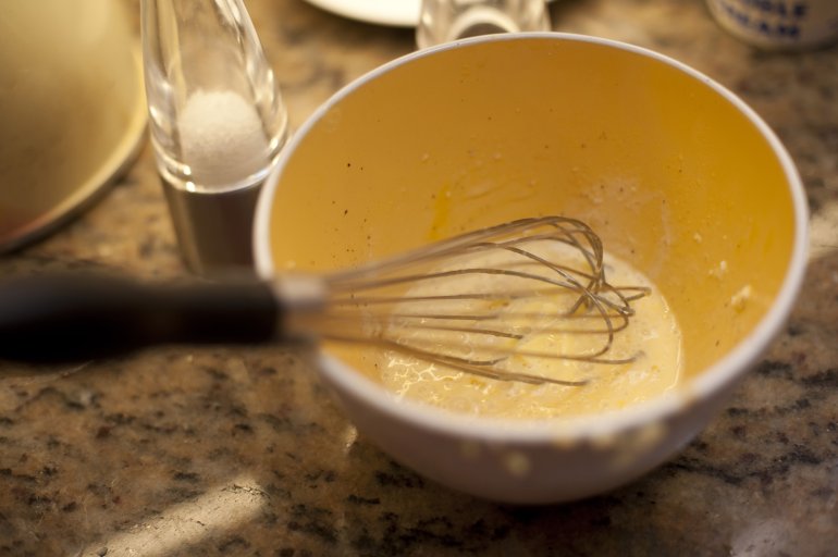 Metal whisk in a mixing bowl with whisked egg on a kitchen table reading for baking