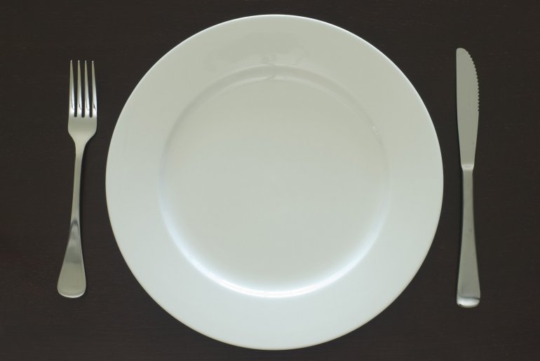 Plain white clean empty dinner plate and cutlery place setting on a black table viewed from above