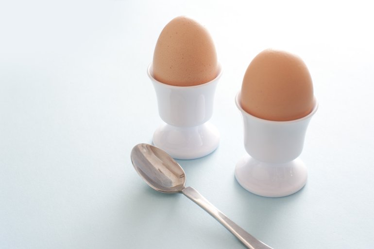 Two fresh boiled eggs for breakfast served in white ceramic egg cups with a spoon over blue with copy space