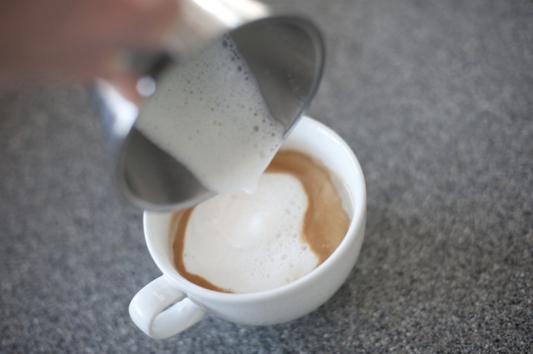 Adding steamed frothy milk to a cup of freshly brewed cappuccino coffee, closeup of a hand pouring the milk from a metal jug, focus to the coffee