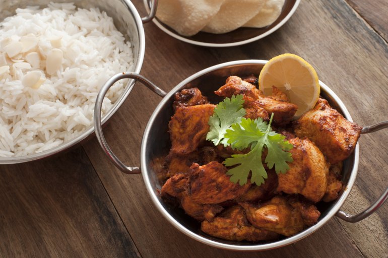 Small silver pot of tandoori chicken with lemon slice and cilantro besides white rice and crispy papadums