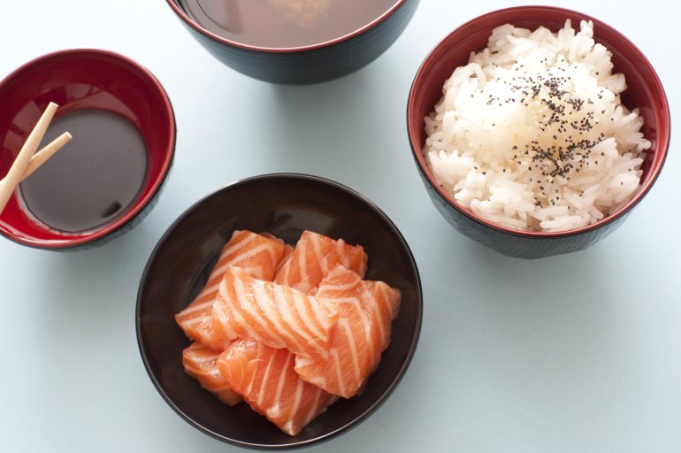 Overhead view of blue table set with asian meal made of salmon chunks, steamed rice and soy sauce in round dark colored bowls