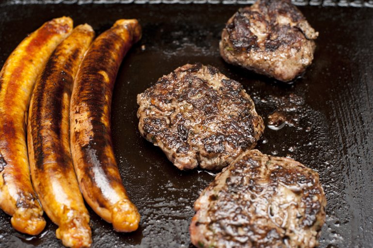 Greasy barbecue griddle with three grilled sausages and three portions if steak cooking on a greasy surface, high angle view