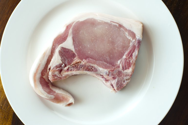 Uncooked fatty pork chop or cutlet on a white plate ready for cooking, overhead close up view