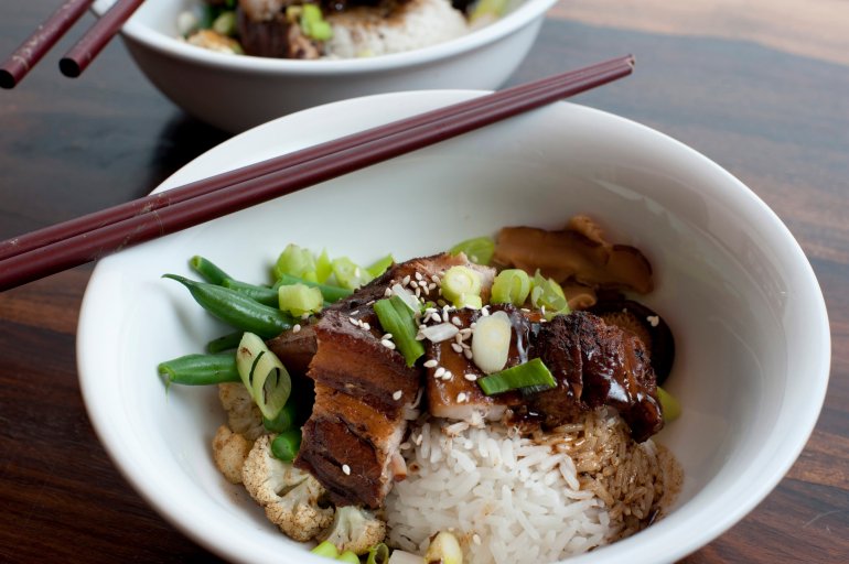 Chinese red pork served in a bowl with rice, cauliflower and beans with chopsticks resting on the rim