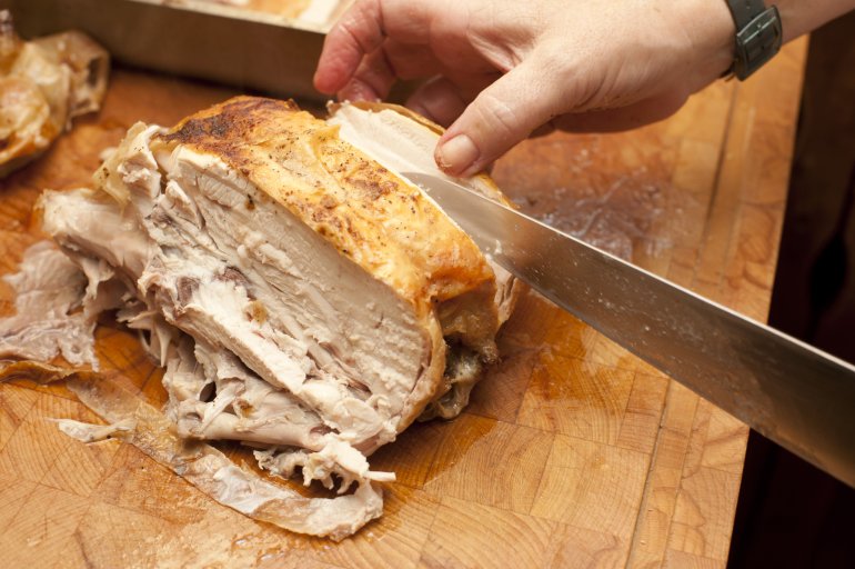 Man carving a half roast chicken on a wooden counter top in the kitchen with a large carving knife