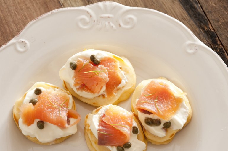Capers and raw fish blini appetizers on white porcelain decorative plate over wooden table