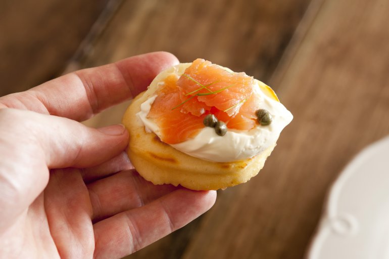 First person view of person eating a blini appetizer with cream, pink salmon and capers in hand