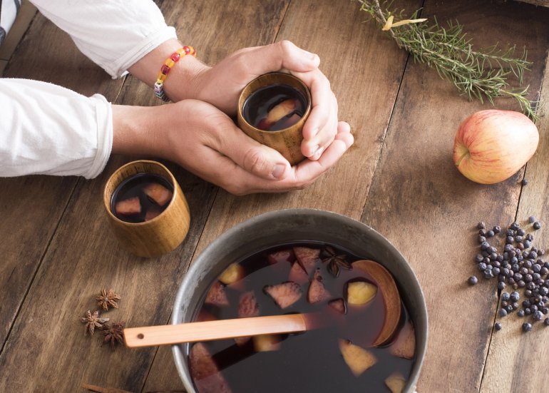 Person making mulled red wine in a bowl with fruit, herbs and spices with ingredients and two full mugs to celebrate the holiday season