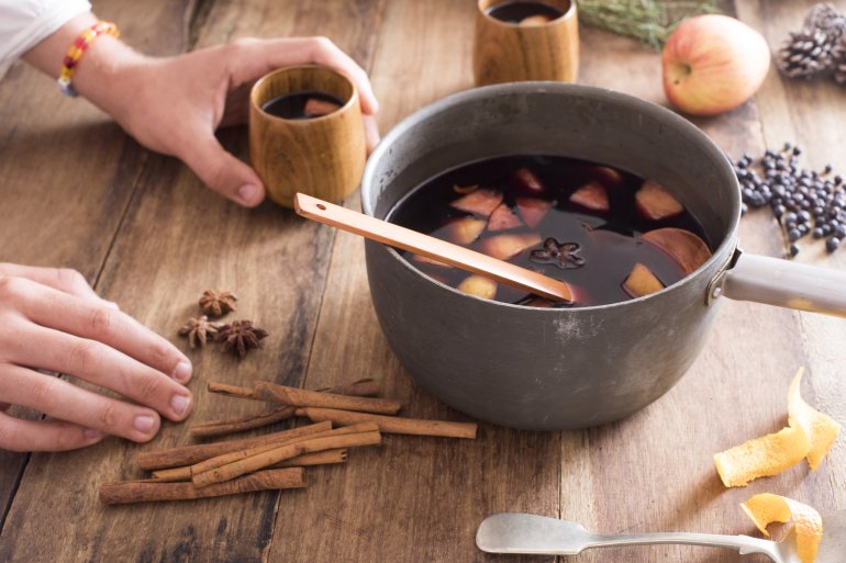 Person serving hot mulled wine with cinnamon, spices and chopped citrus fruit from a bowl on a wooden table with scattered ingredients
