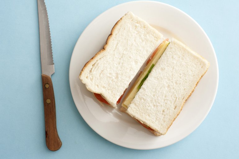 Top down view on white bread sandwich filled with cheese and vegetables over single round white plate and knife on blue table
