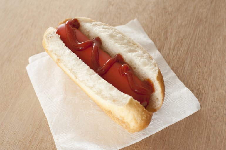 First person perspective view of large red hot dog with ketchup on square white napkin over light brown table