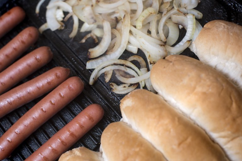 Cooking hot dog ingredients on a griddle with a row of smoked Wiener or Frankfurter sausages, diced onion and fresh white bread rolls