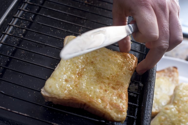 Man holding a knife in his hand removing a slice of golden toasted cheese from a griddle in the kitchen for a tasty lunchtime snack