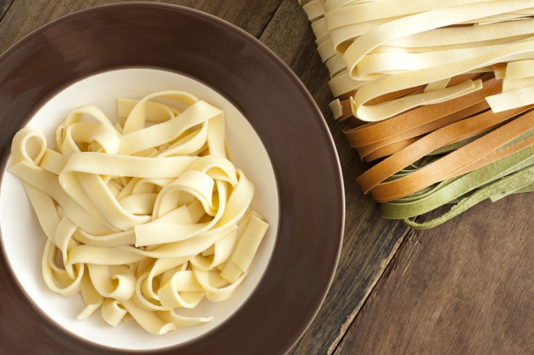 Cooked and dried Italian tagliatelle pasta viewed from above with a serving of plain boiled pasta in a bowl alongside white, brown and green dried ribbons on a wooden table