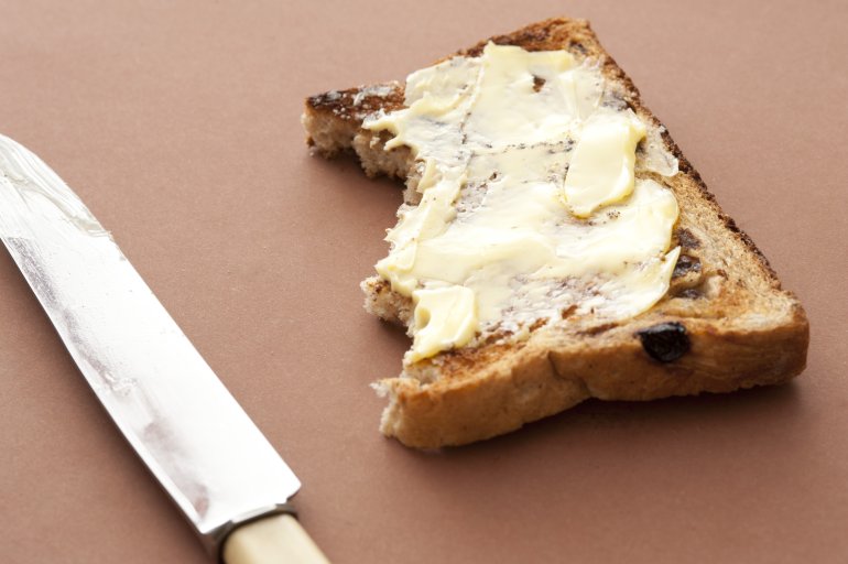 Half eaten slice of buttered raisin bread with bite marks on a brown background alongside a knife