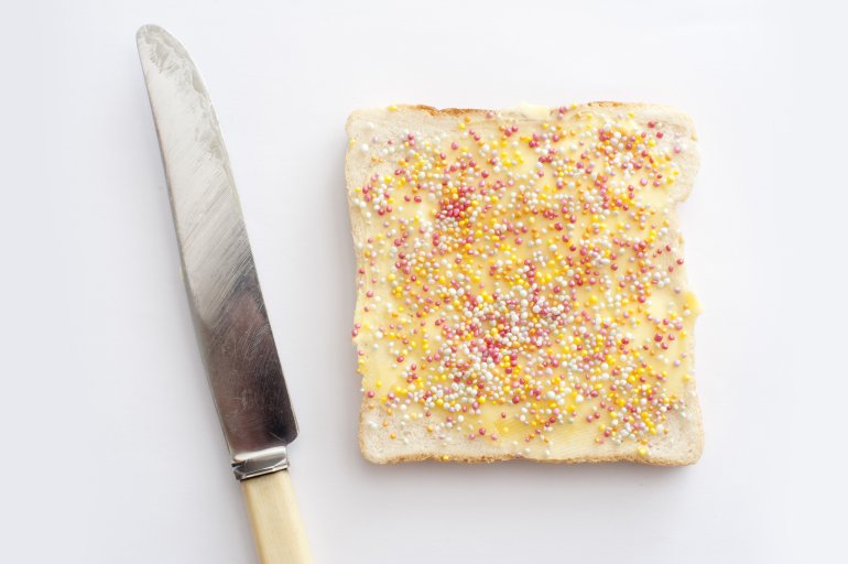 Single slice of party fairy bread with a topping of colorful candy sprinkles viewed from above alongside a knife on a white background