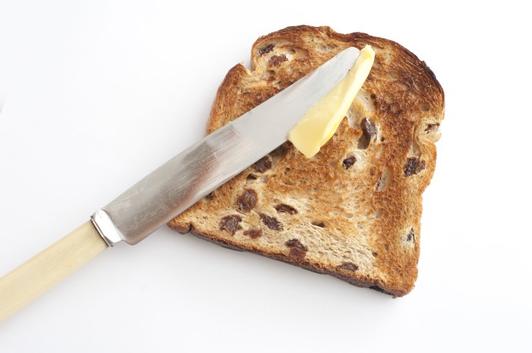 Spreading butter on a slice of hot fresh fruity toast with raisins using a bone handled knife, high angle view on white