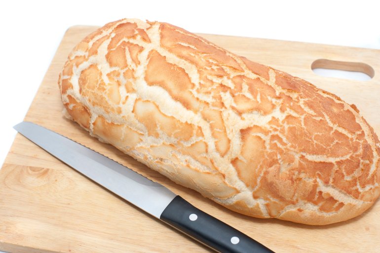 Loaf of freshly baked crusty white bread on a wooden bread board in the kitchen with a bread knife, white background