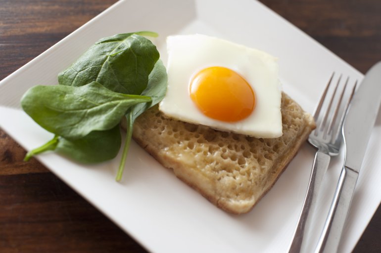 Close up of square egg sunny side up on a crumpet besides fresh spinach leaves and utensils on a white plate