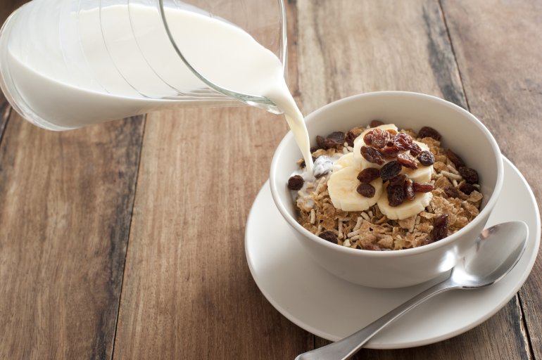 Milk being poured into round bowl filled with muesli, raisins and banana slices beside metal spoon on wood surface