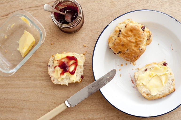 First person perspective view looking downward at two baked scones with butter, fruit jam and knife over wooden table