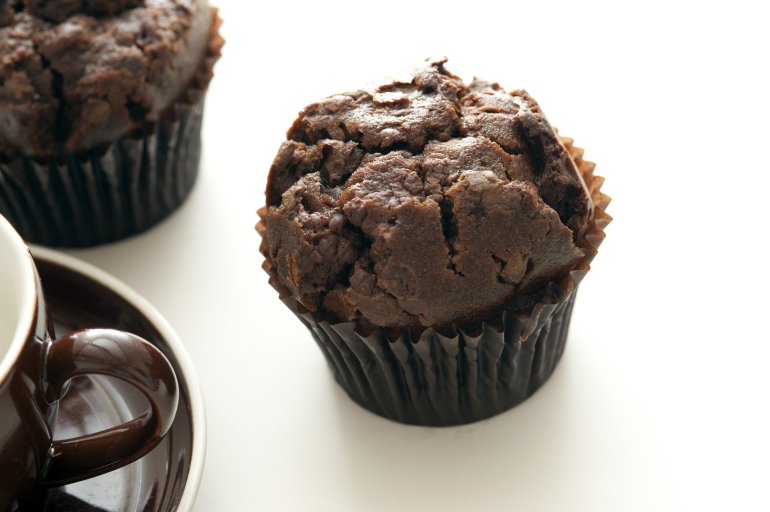 Tasty freshly baked double chocolate muffins next to cropped coffee cup on white table