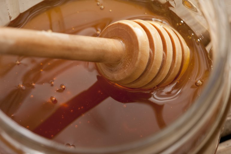 Dipping into a glass jar of fresh honey with a wooden dip stick to drizzle it over the top of food, close up view of the utensil and surface of the honey