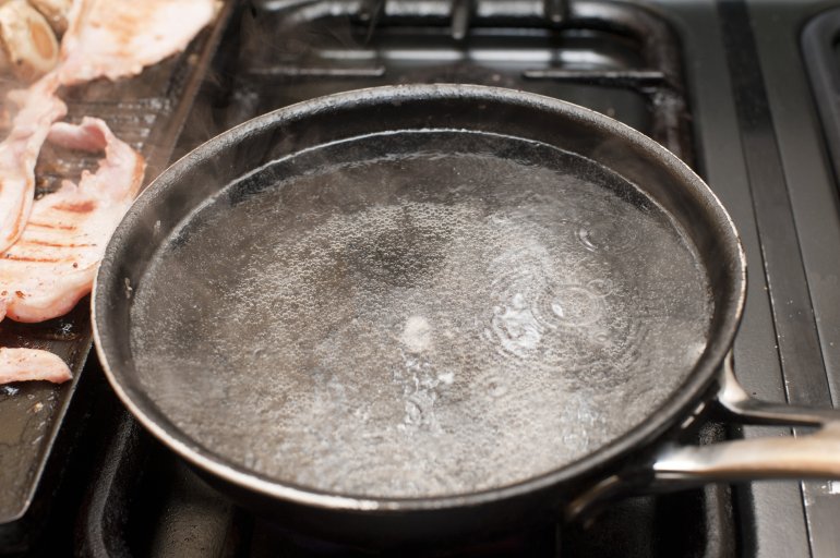 Water in non stick frying pan boils ready for poacking eggds and emits steam while meat cooks on grill nearby