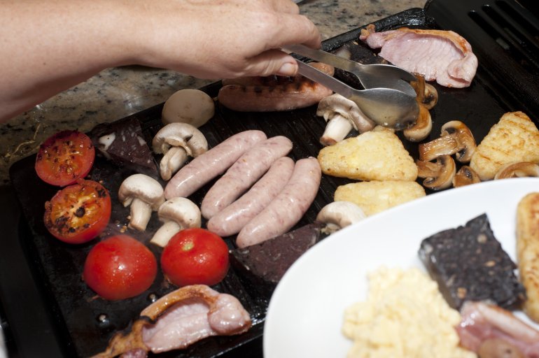 Man preparing a wholesome breakfast serving bacon, black pudding, mushrooms sausages and tomato onto a plate from a hot griddle