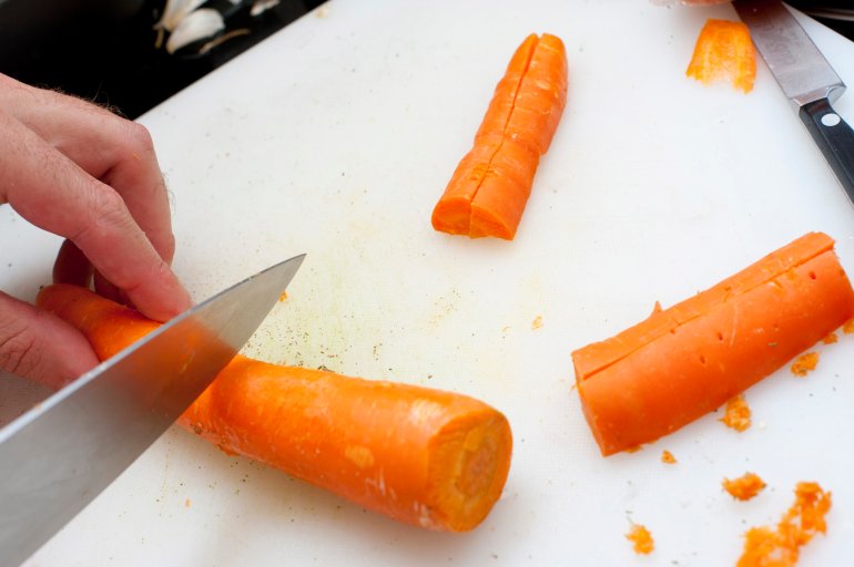 Man cutting fresh whole carrots which he has scraped and cleaned using a large chefs knife as he prepares the vegetables for a meal