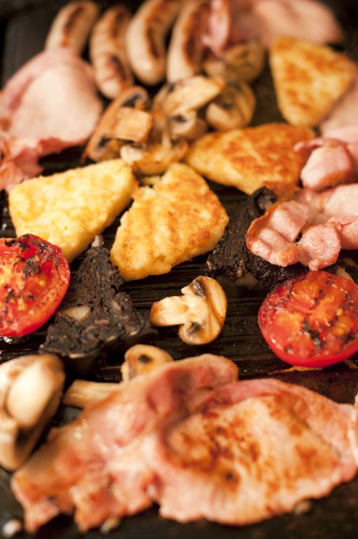 Close up view of grilled tomato and mushroom cooking besides four sausage links and other meats