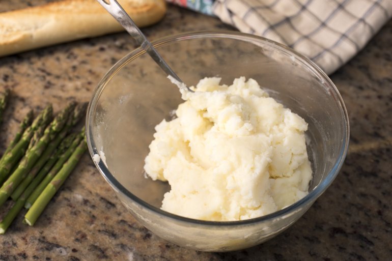 Bowl of freshly mashed cooked potato standing alongside fresh asparagus tips on a marble kitchen counter