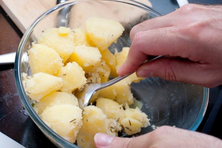Man preparing parboiled cleaned and diced potatoes in a glass bowl stirring them with a spoon to season them during cooking