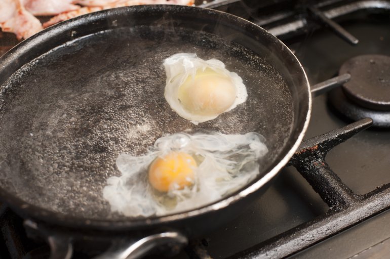Two eggs being poached in boiling water on a gas range beside cuts of meat